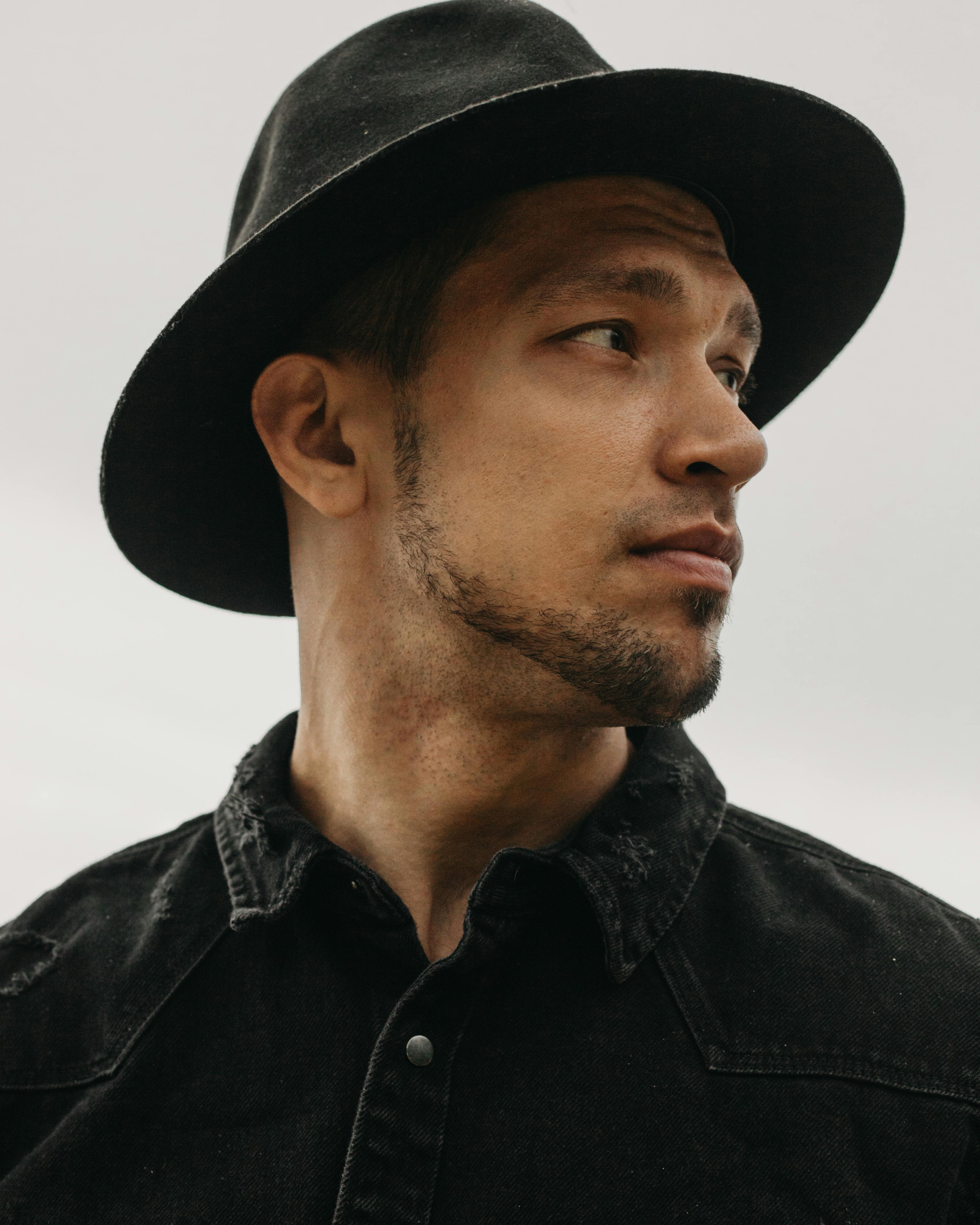 Handsome bearded male in black denim shirt wearing trendy headwear and looking into distance on white background in light room