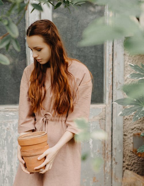 Peaceful female with red hair looking down while standing with flowerpots in hands near shabby wooden door and blurred green leaves