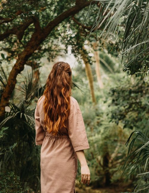 Back view of unrecognizable female with red hair strolling among tall lush trees with green leaves in garden on summer day