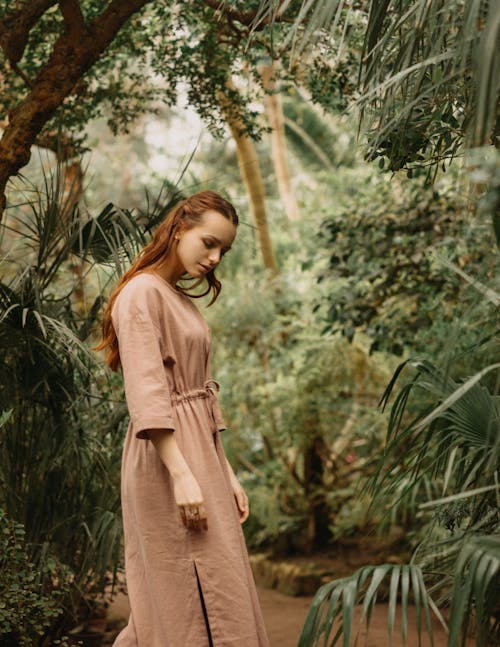 Side view of dreamy female in stylish clothes looking down while standing among tall lush green plants in garden on summer day