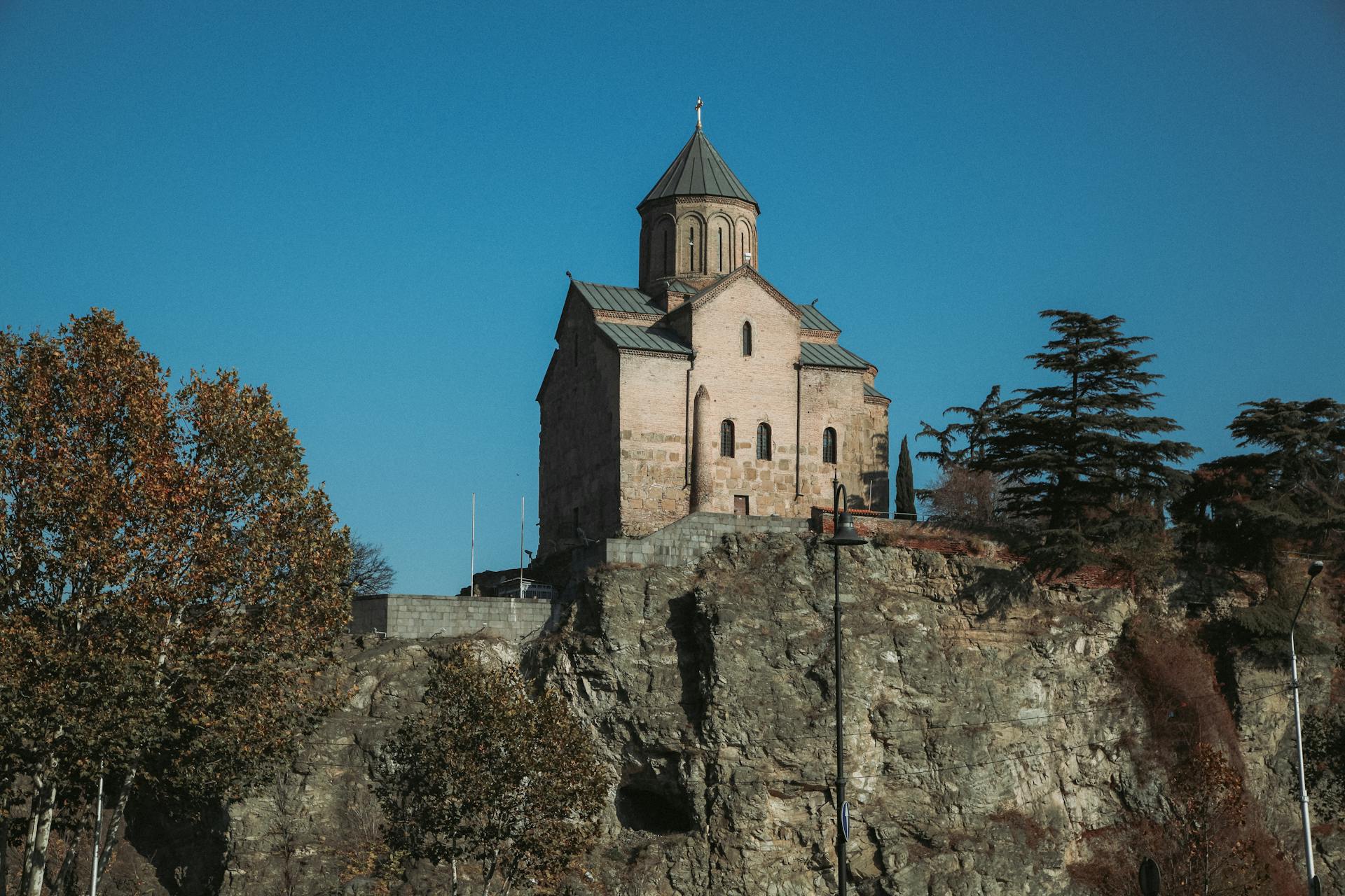 Metekhi Church perched on a cliff in Tbilisi against a clear blue sky, surrounded by trees.