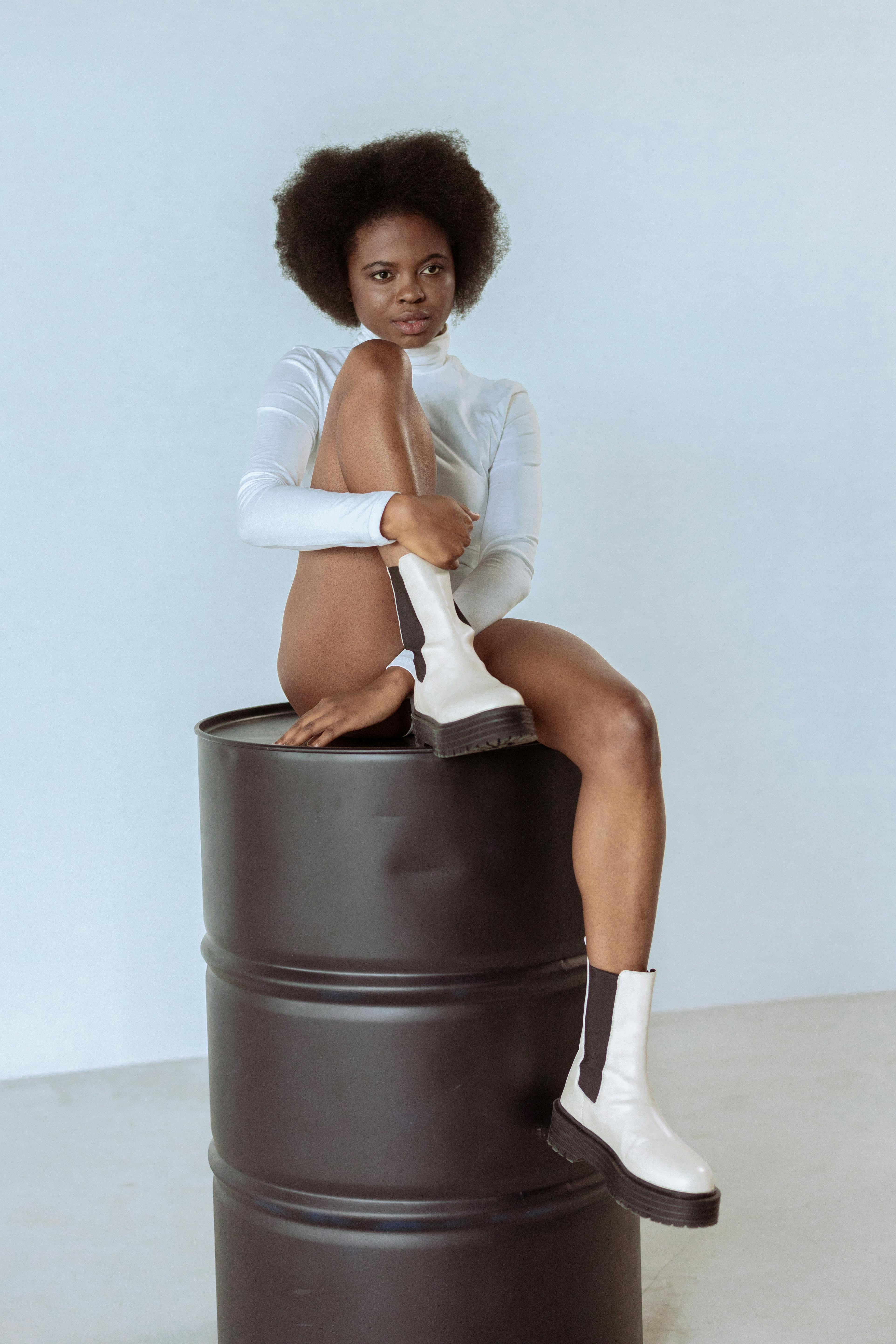Stylish photoshoot of a woman in a bodysuit posing on a barrel in a studio setting.