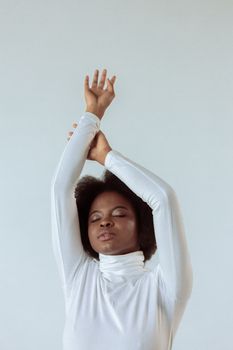 Studio portrait of a beautiful woman in white attire with afro hair, arms gracefully raised, on a plain background.