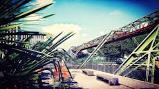 在城市环境中，可以看见一座现代钢桥，桥边有长凳和树木，天空晴朗