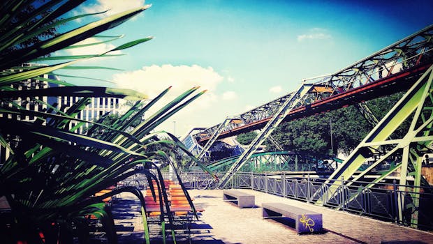 在城市环境中，可以看见一座现代钢桥，桥边有长凳和树木，天空晴朗