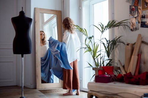 Woman in White Blouse Looking at Her Reflection on the Mirror while Holding a Blue Fabric