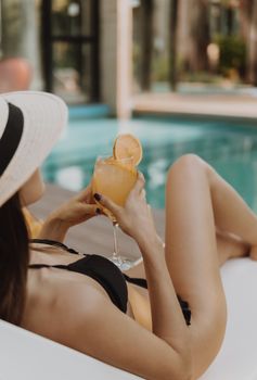 Woman in bikini relaxing by the pool, holding an orange juice, capturing a serene summer moment.