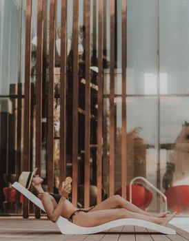 Woman in bikini relaxing on a sun lounger, enjoying a drink by the poolside at a luxury resort.