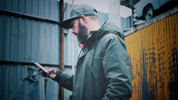 Side view of a man with a beard and cap using a smartphone outdoors.