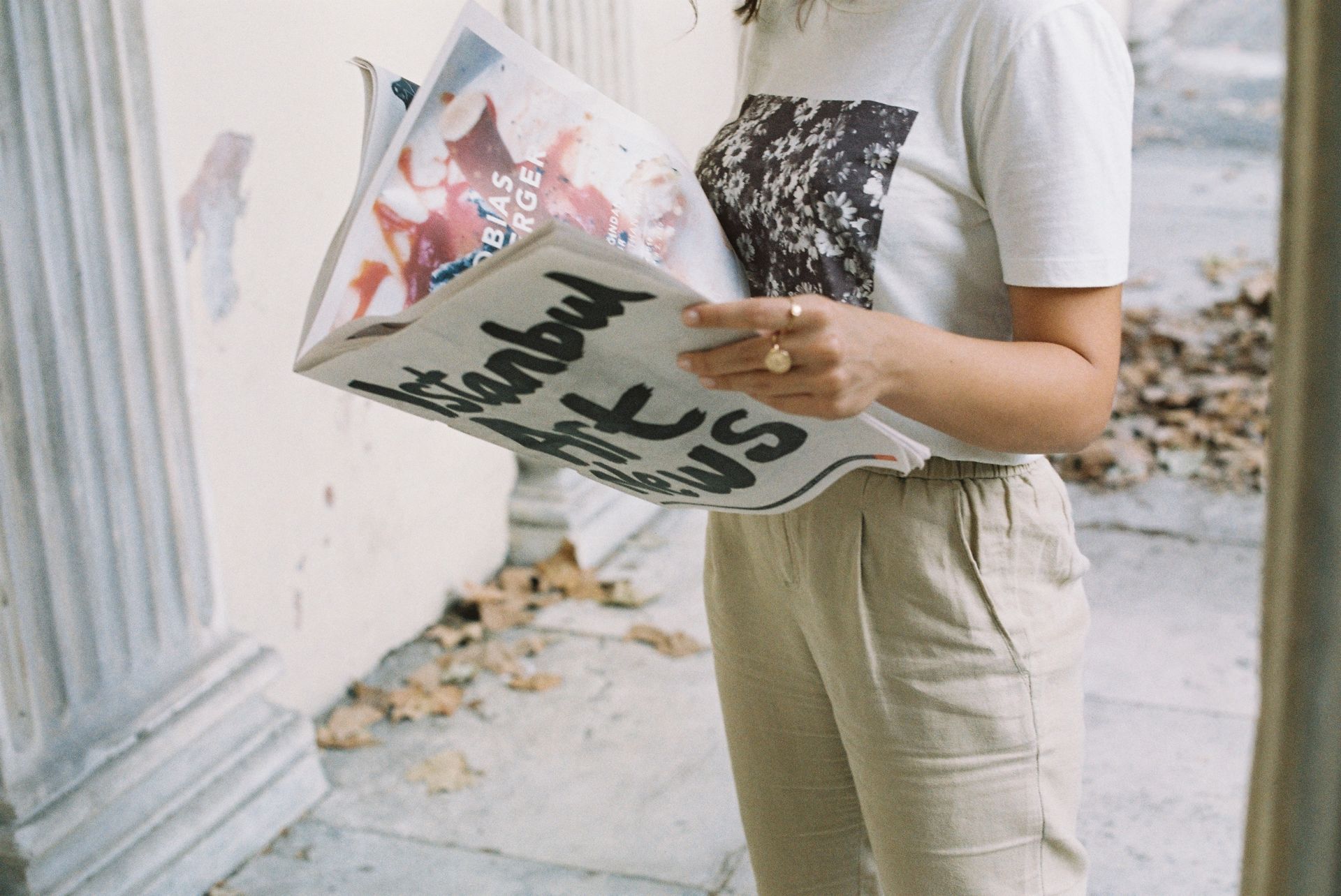A person reading the Istanbul Art News in an urban outdoor setting with fallen leaves and columns.
