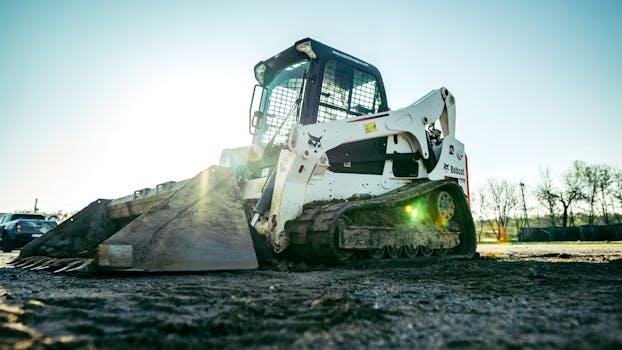 A skid steer loader on a construction site with bright sunlight in the background.