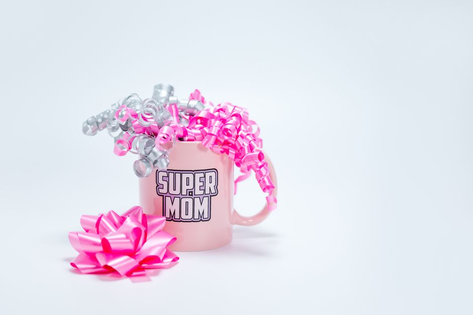 Uma caneca Super Mãe rosa adornada com fitas enroladas rosas e prateadas sobre um fundo branco, perfeita para o Dia das Mães.
