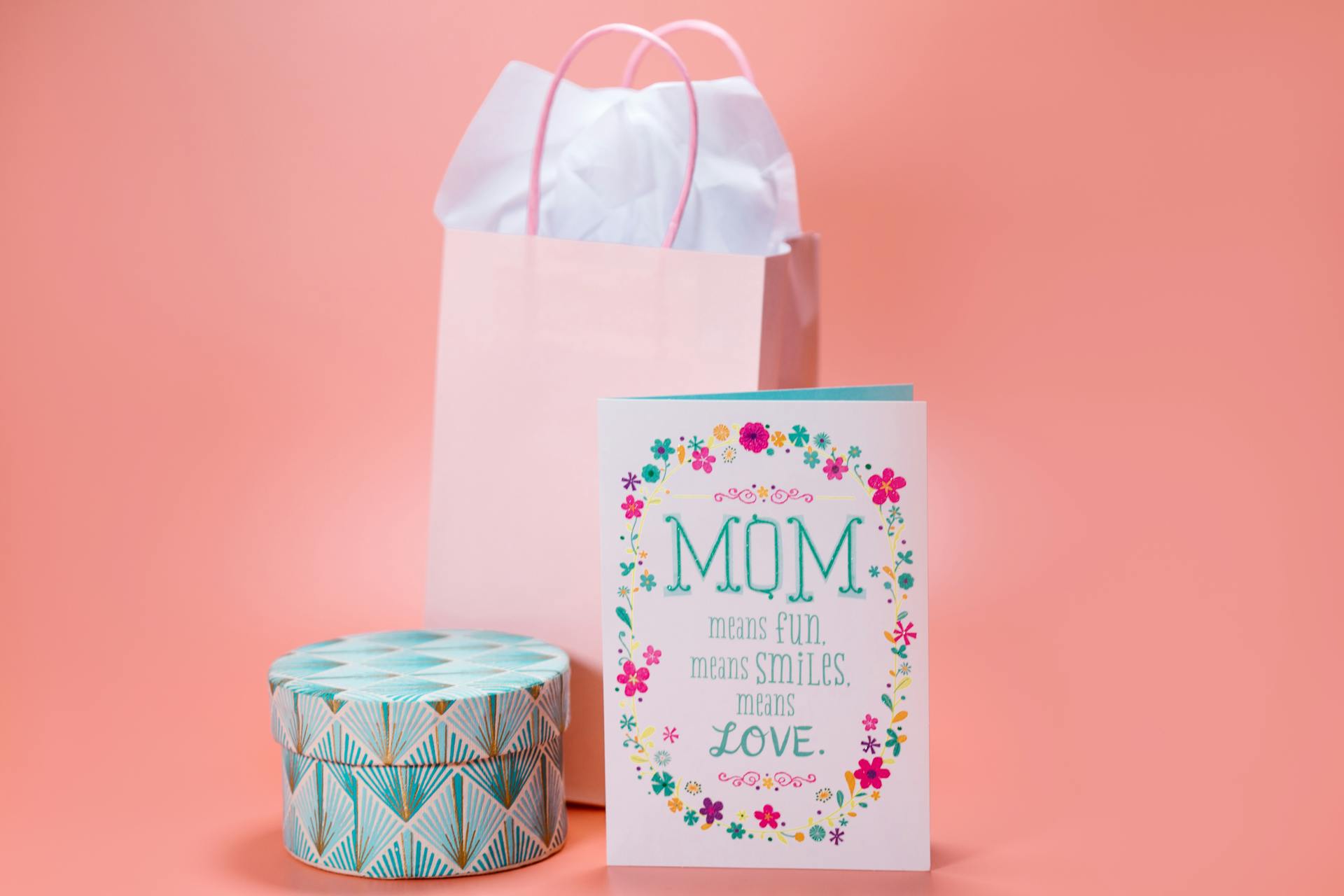 Elegant Mother's Day greeting card with gift bag and decorative box on pastel pink background.