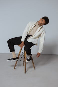 Portrait of a fashionable man with casual outfit posing on a wooden stool in a minimalist studio.