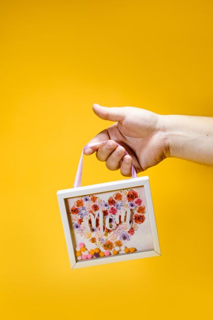 A hand holds a floral picture frame spelling 'Mom' against a bright yellow background.
