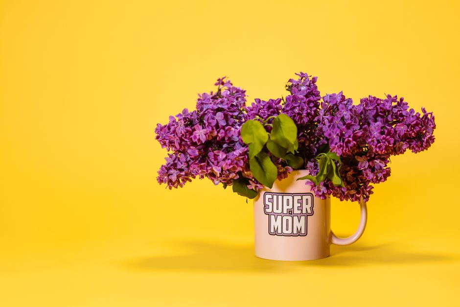 Uma exibição vibrante de lilases roxos em uma caneca 'Super Mãe' contra um fundo amarelo brilhante, perfeita para temas de maternidade.