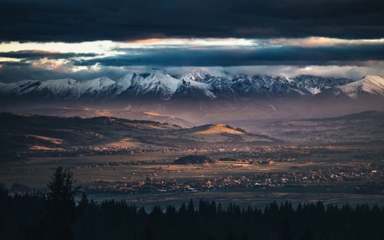 Snow-capped mountains under heavy clouds with sunlit valley settlements