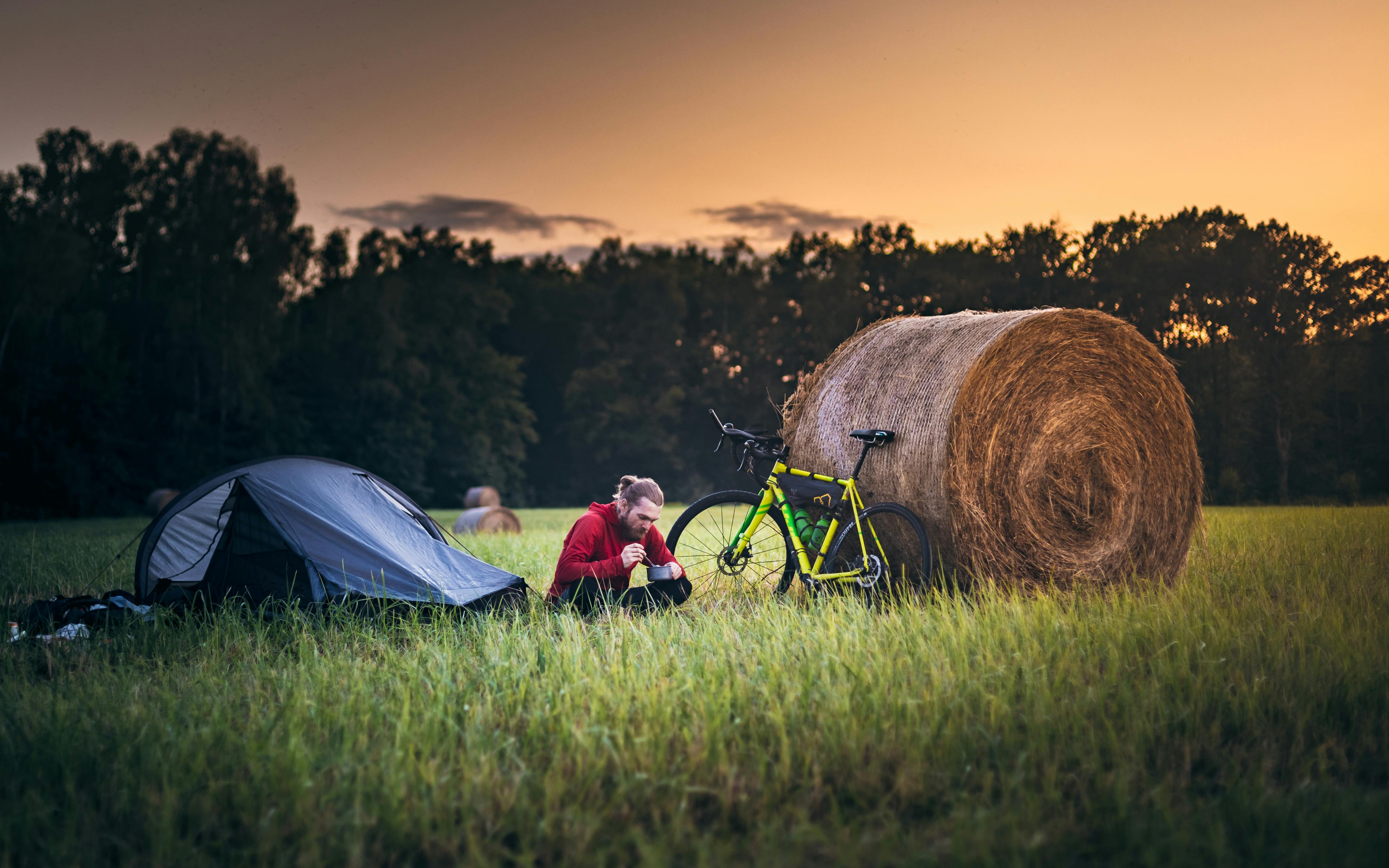 A man camping with a bicycle and tent near a hay bale at sunset in the Polish countryside.