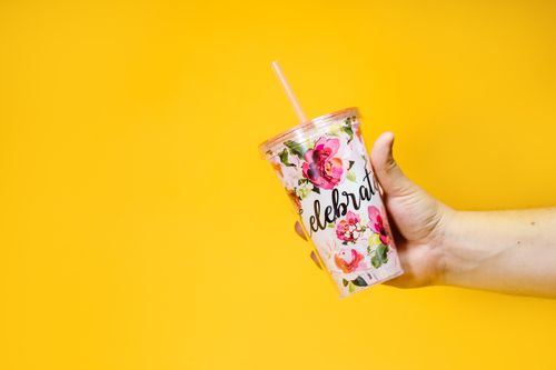 Free Close-up of a hand holding a floral tumbler with straw on a vibrant yellow background. Stock Photo