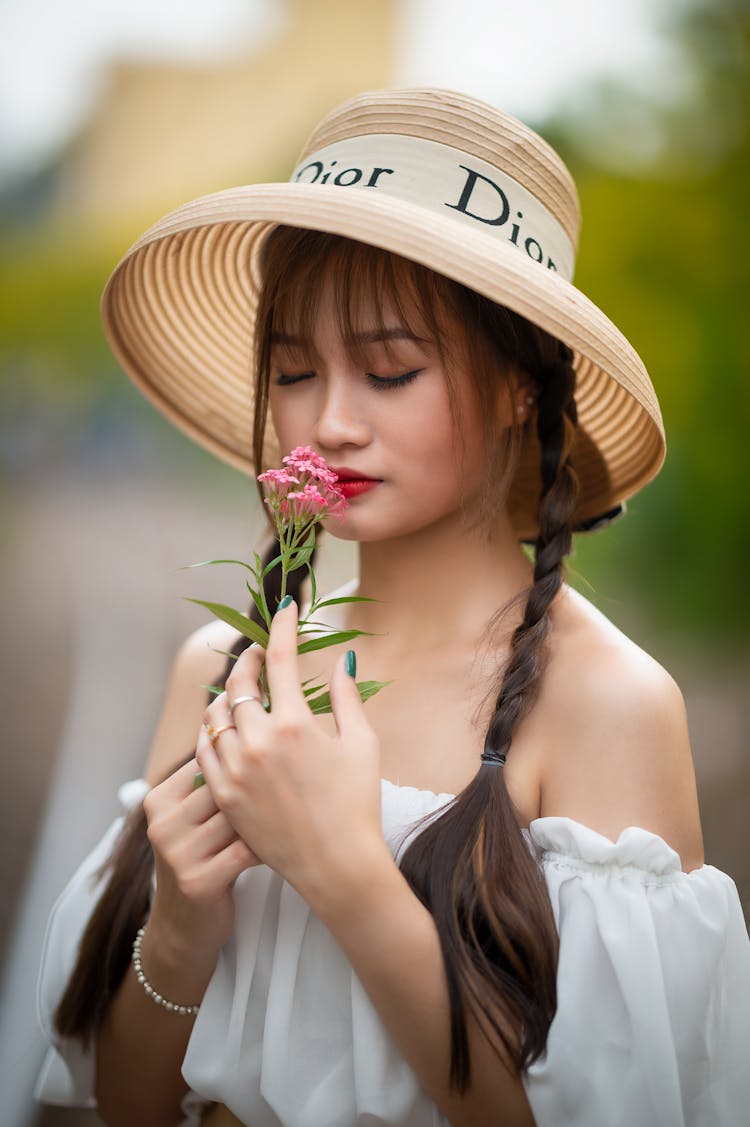 A Pretty Woman In White Off Shoulder Top And Dior Sun Hat Smelling Flowers