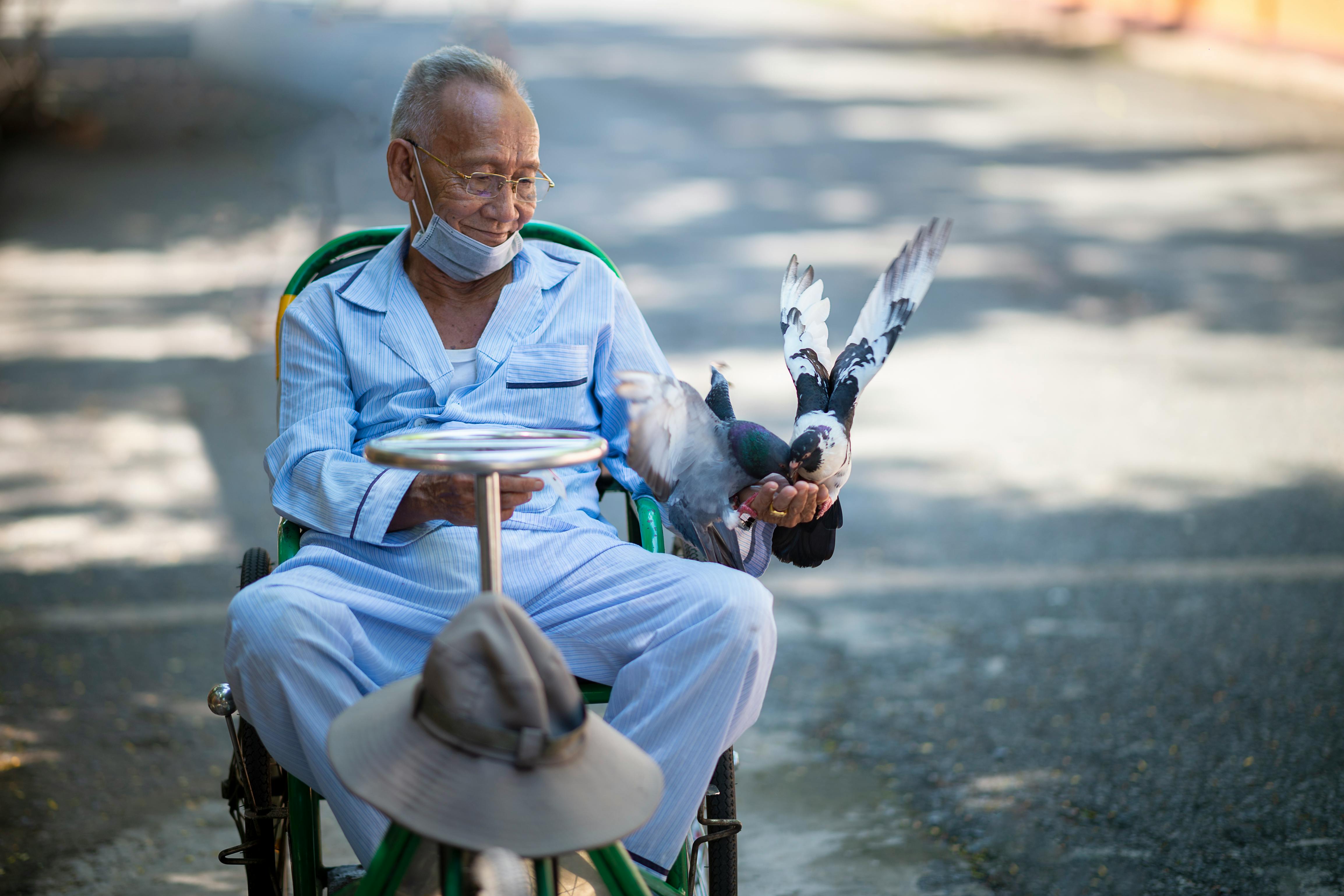 Senior man in wheelchair joyful while feeding birds, wearing pajamas and face mask outdoors.