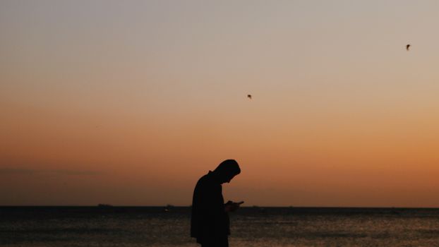 Side view silhouette of anonymous male on coastline texting on mobile phone against sky with picturesque sunset