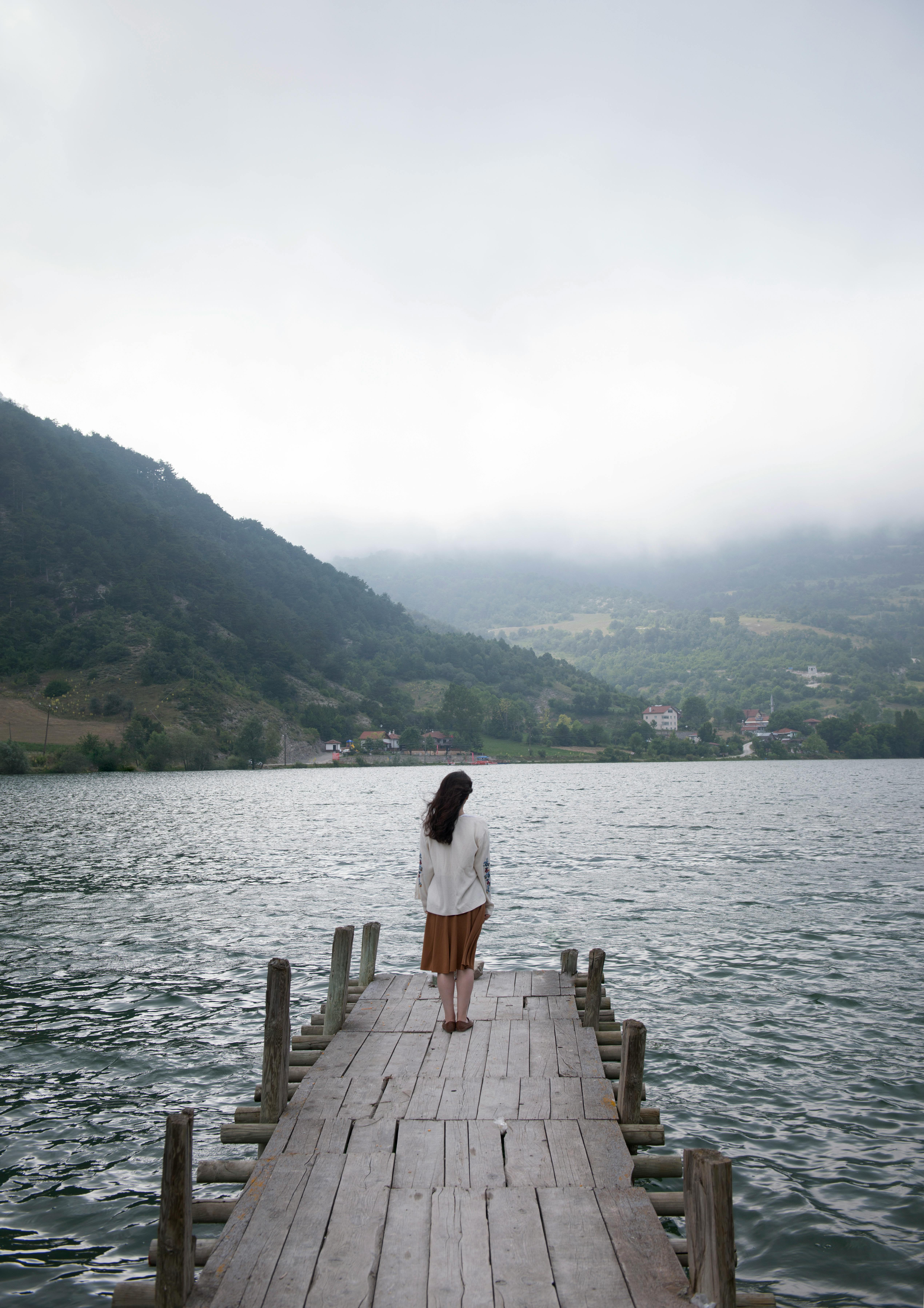 Back view of distant faceless female standing on wooden quay near rippling water against grassy hills and fog in nature