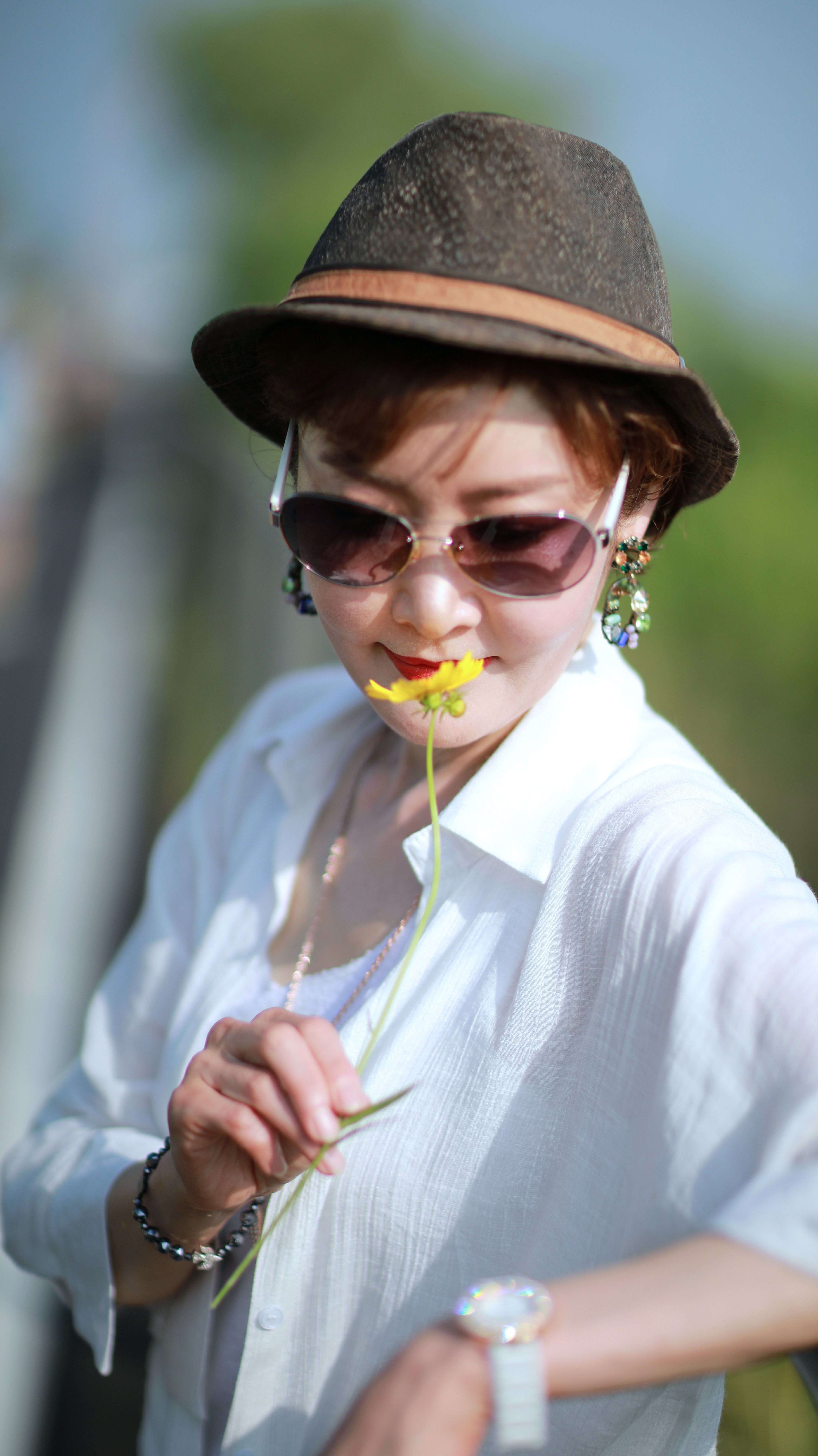 Woman in hat and sunglasses appreciating a yellow flower outdoors on a sunny day.