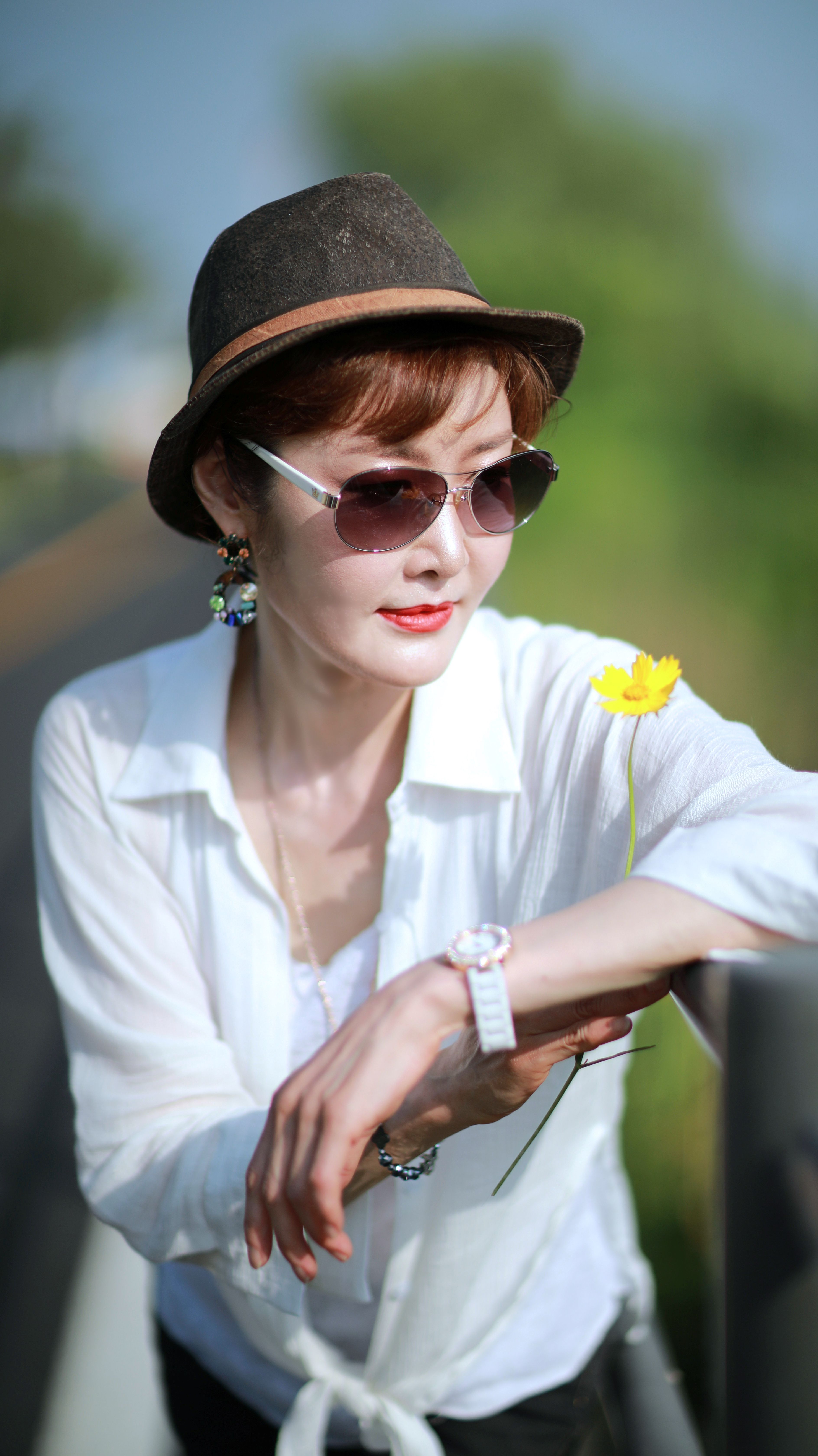 Woman wearing sunglasses and hat holding yellow flower outdoors in bright daylight.