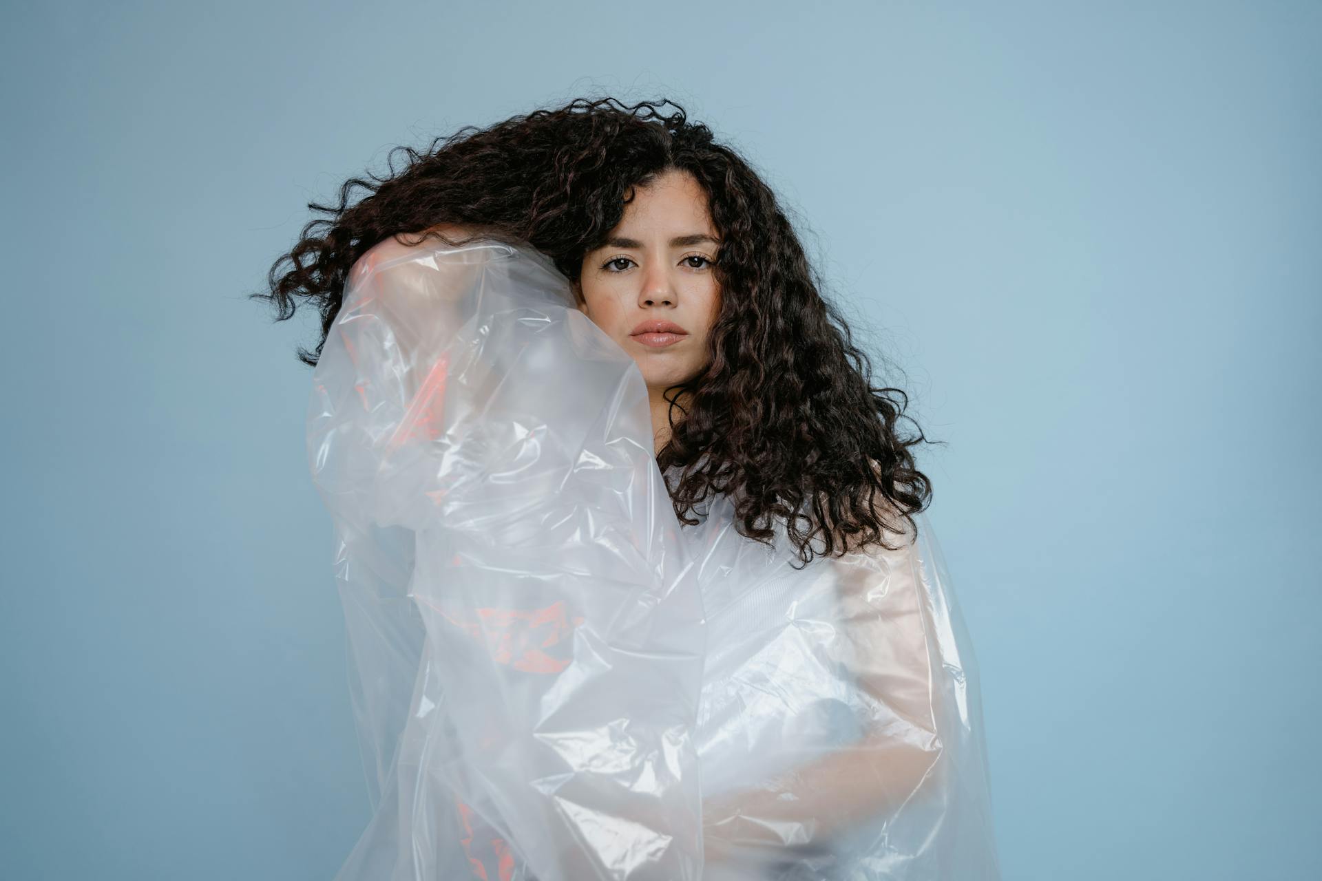 A woman with curly hair wrapped in transparent plastic against a blue background.