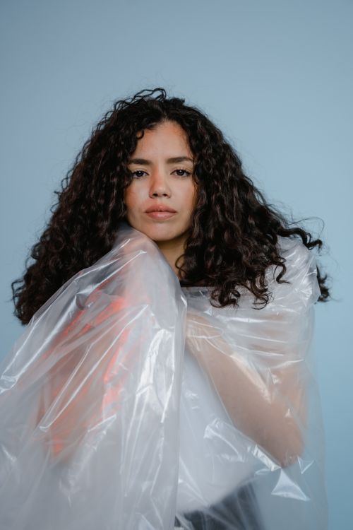 Free Artistic portrait of a woman with curly hair wearing a translucent plastic wrap against a blue background. Stock Photo