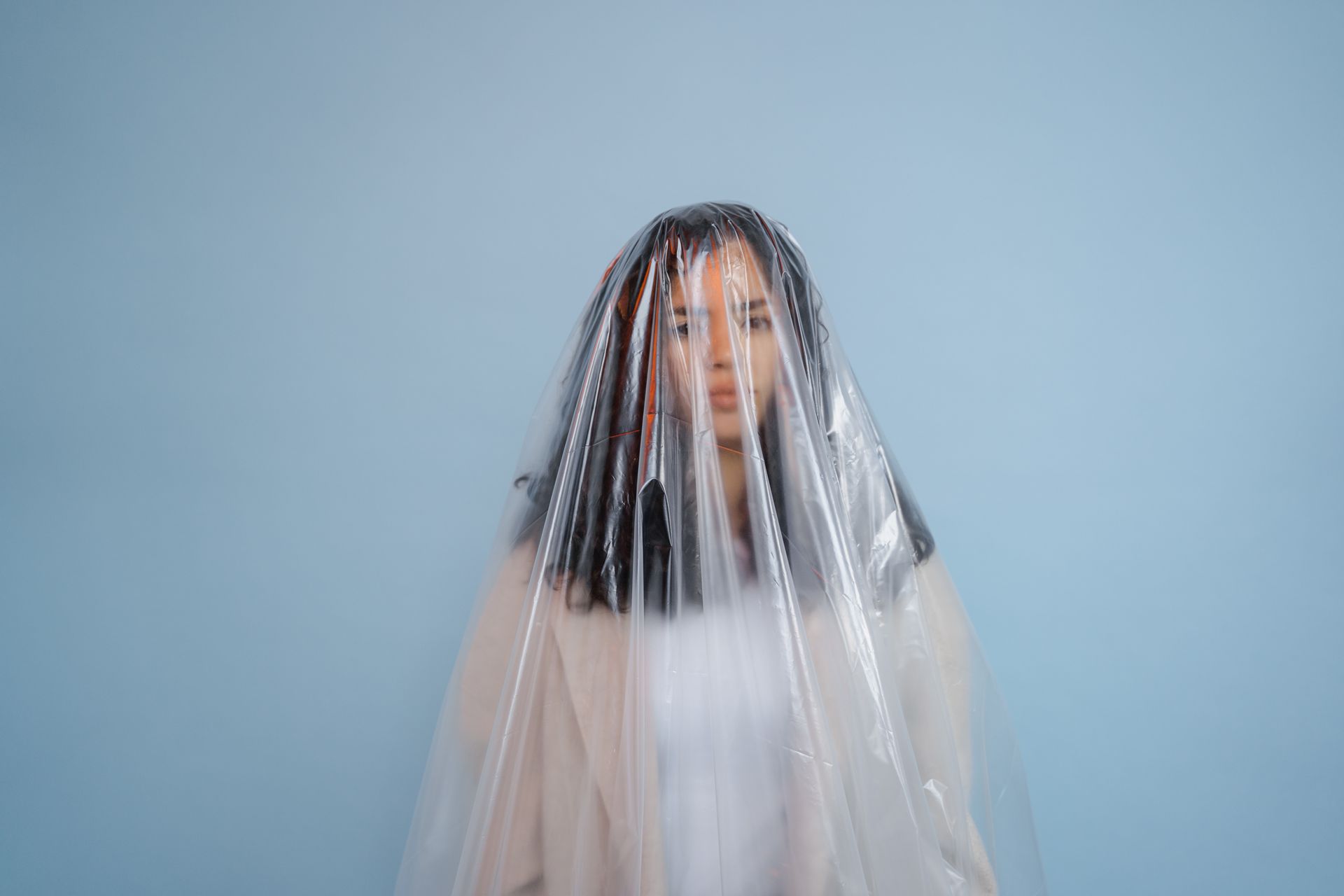 Portrait of a woman draped in plastic, isolated against a soft blue background, evoking concepts of concealment and introspection.