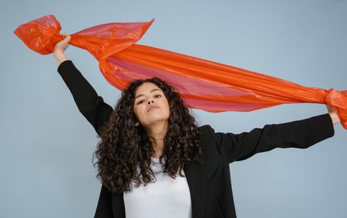 Free Confident young woman holding a vibrant orange sheet against a light blue backdrop. Stock Photo
