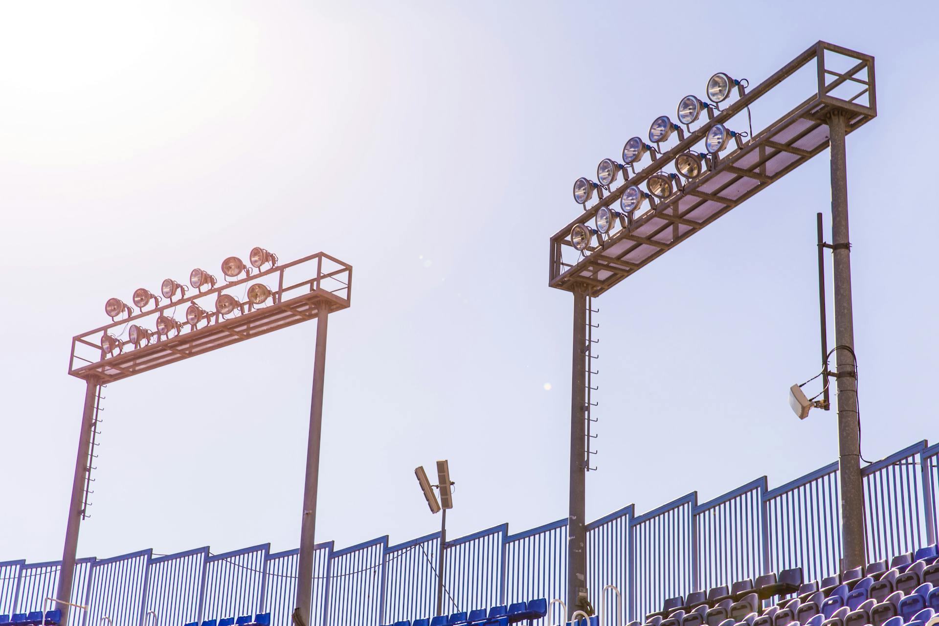 Bright stadium spotlights under a clear blue sky by empty seats, symbolizing sports events.