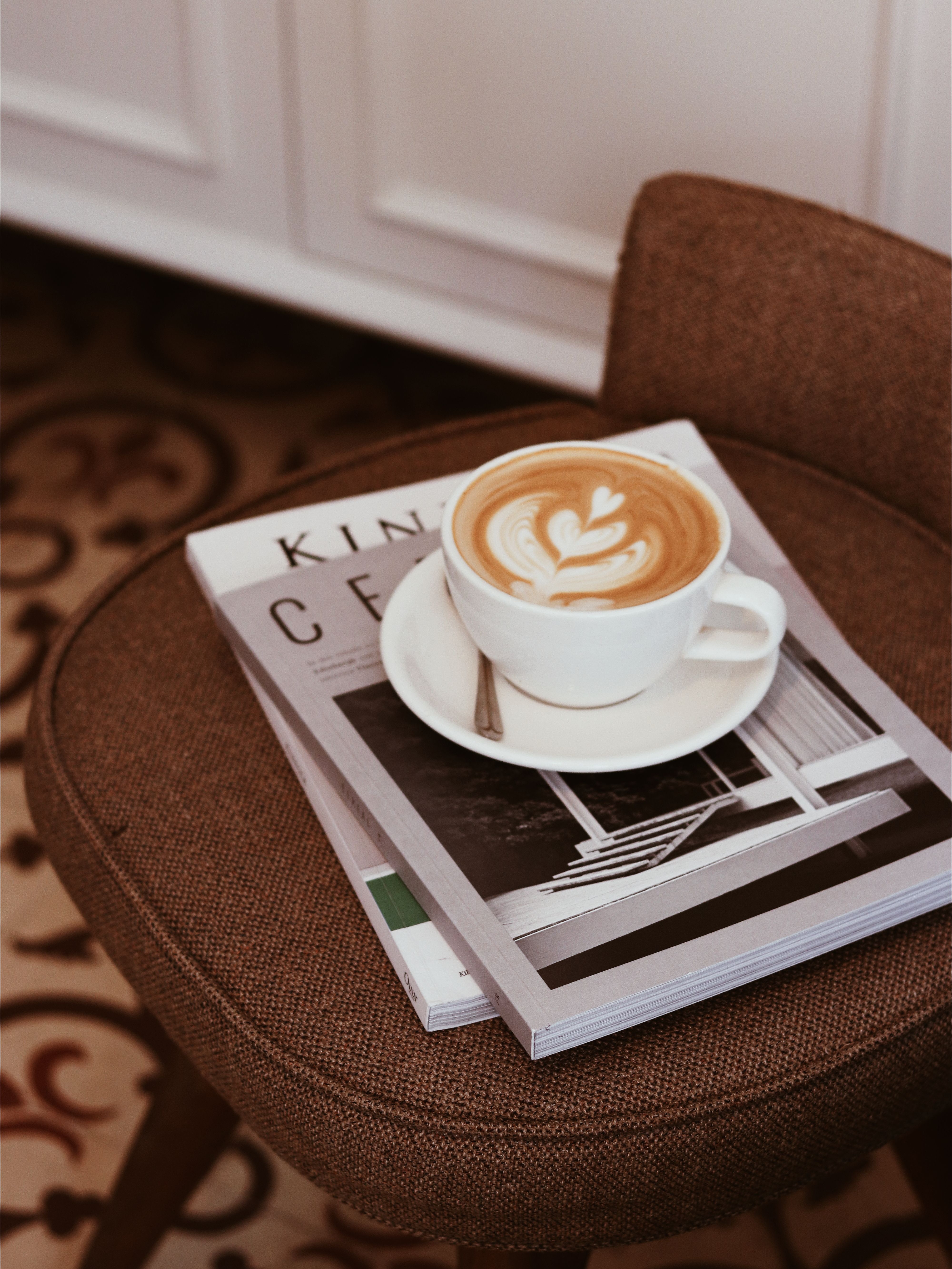 A cup of coffee with latte art on a magazine, set on a brown chair indoors, perfect for cozy moments.
