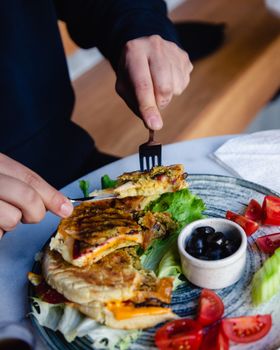Close-up of a person slicing savory toast with vegetables and a side of olives on a ceramic plate.
