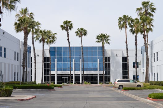 Contemporary glass office building with palm trees and a parked car in daylight.