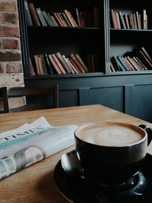 Free A warm setting with a coffee cup, bookshelves, and newspaper on a wooden table. Stock Photo
