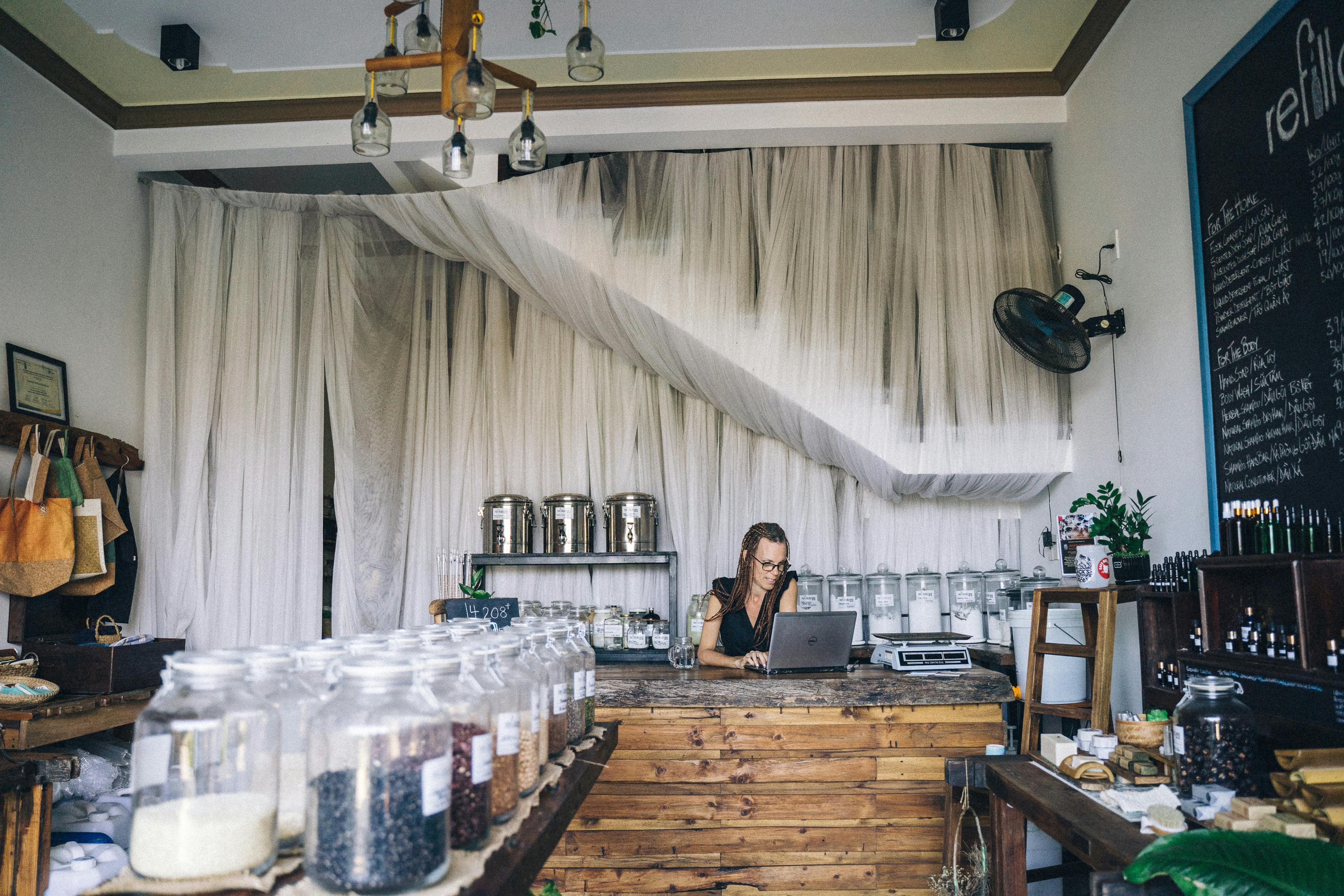 Woman working on laptop in eco-friendly store with glass jars and refillable items.