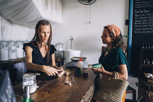 Two women discuss eco-friendly products in a store with refillable glass jars and organic items.