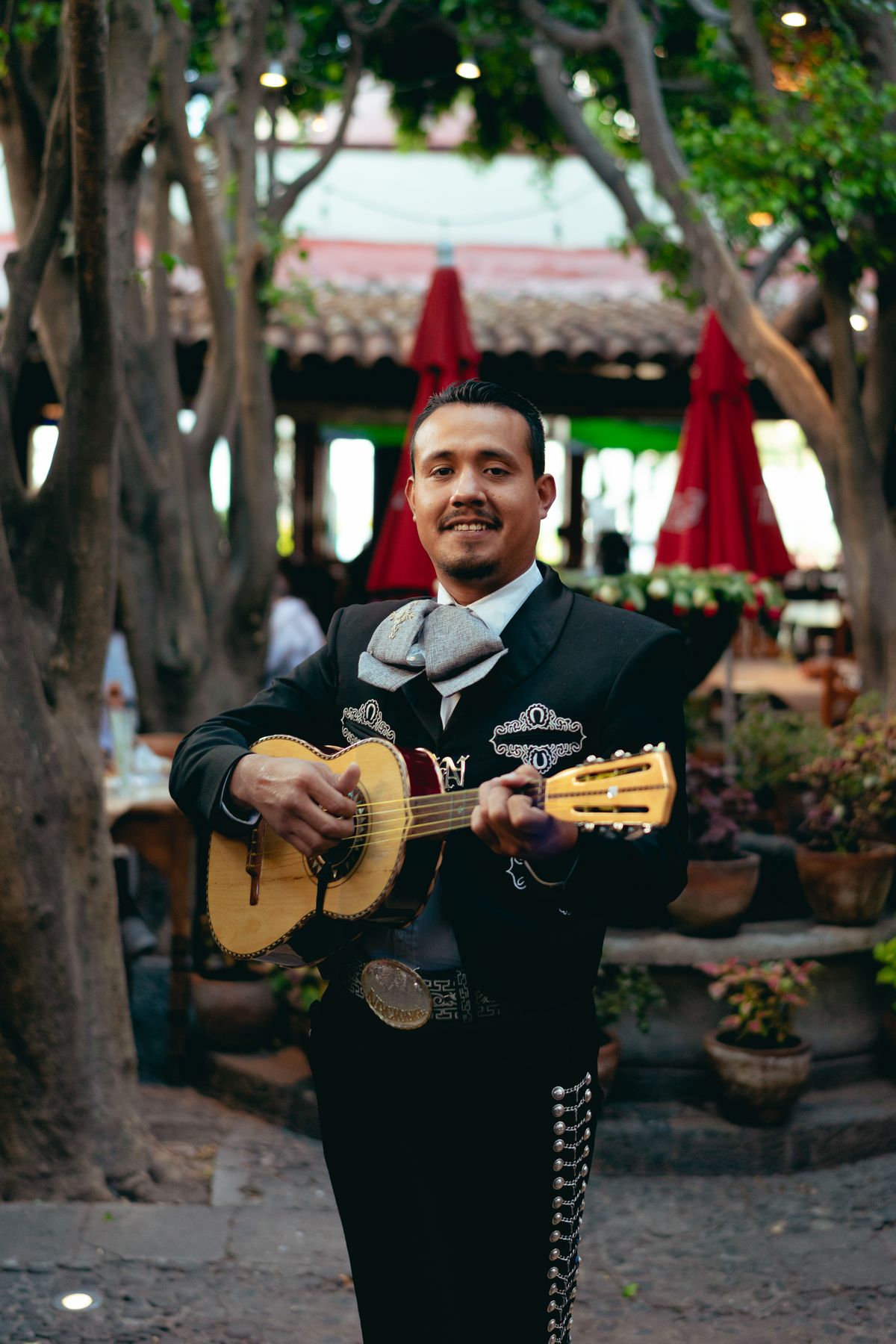 A mariachi musician smiling while playing guitar outdoors, showcasing traditional Mexican attire and culture.