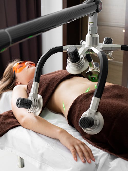 Free Female client in eyewear lying on table under modern laser fat loss machine during liposuction treatment in light medical clinic Stock Photo