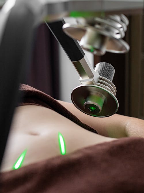 Crop unrecognizable female client with naked belly lying under lasers of fat loss machine with heads during procedure in medical clinic