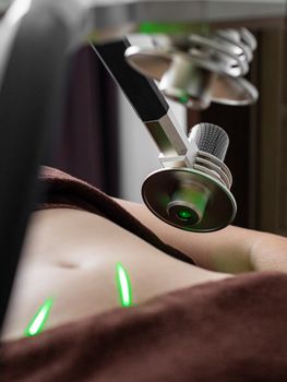 Crop unrecognizable female client with naked belly lying under lasers of fat loss machine with heads during procedure in medical clinic