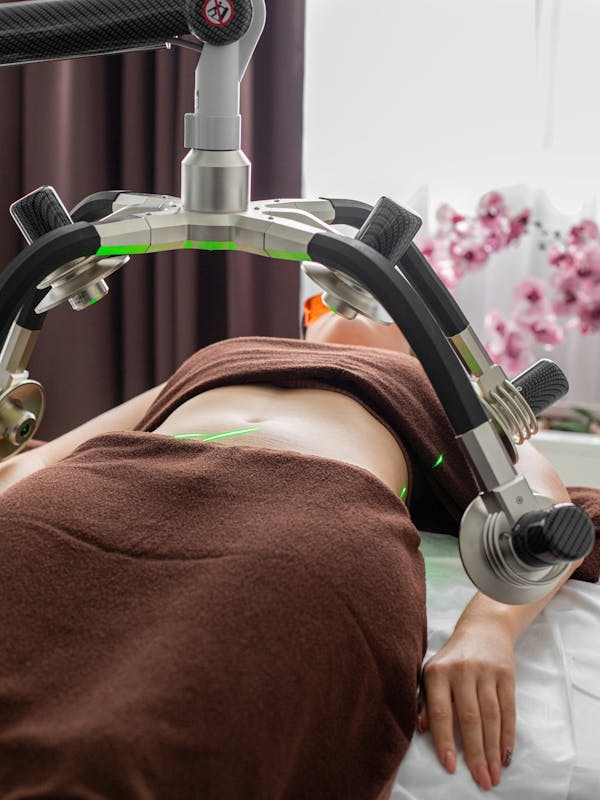 Crop anonymous female patient lying under lasers of fat loss machine on belly during treatment in light modern medical clinic