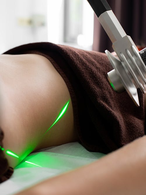Crop anonymous patient with towel lying under green lasers of modern fat loss machine during procedure in light medical clinic