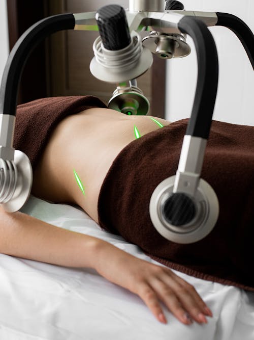 Crop anonymous female client lying under green lasers on belly while receiving fat loss treatment in modern light medical clinic