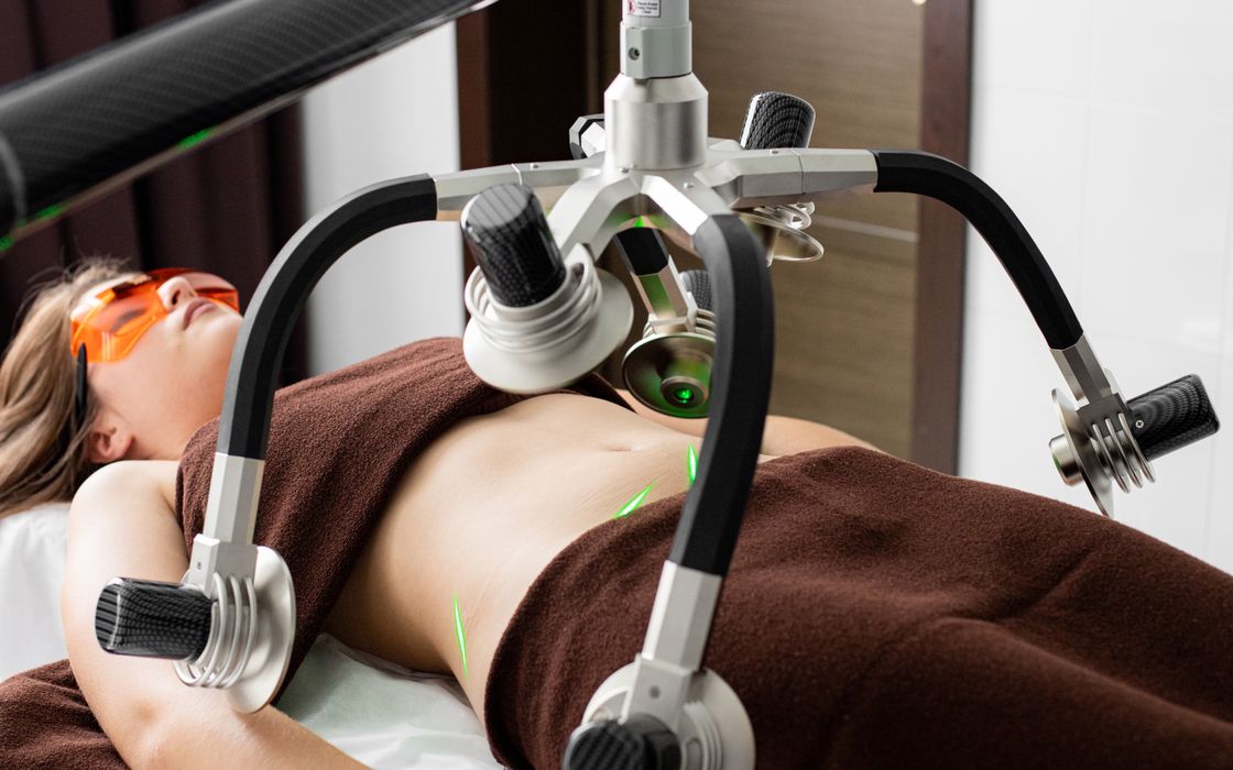 Female client lying on table under lasers of modern weight loss machine on belly during liposuction procedure in medical