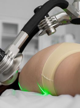 Crop anonymous female patient with lasers on hip and boot lying under weight loss machine during procedure in medical clinic