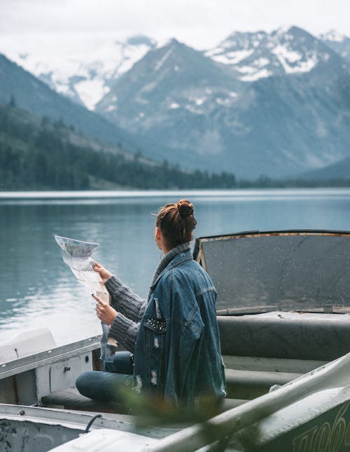 Side view of faceless female in warm clothes sitting with map in boat on lake while admiring view of snowy hills and woods in nature in daytime